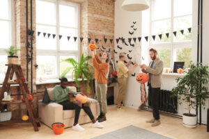 Group of young multi-ethnic friends decorating room for Halloween: girl attaching paper rats on wall, woman and man hanging paper lanterns, black guy carving pumpkin